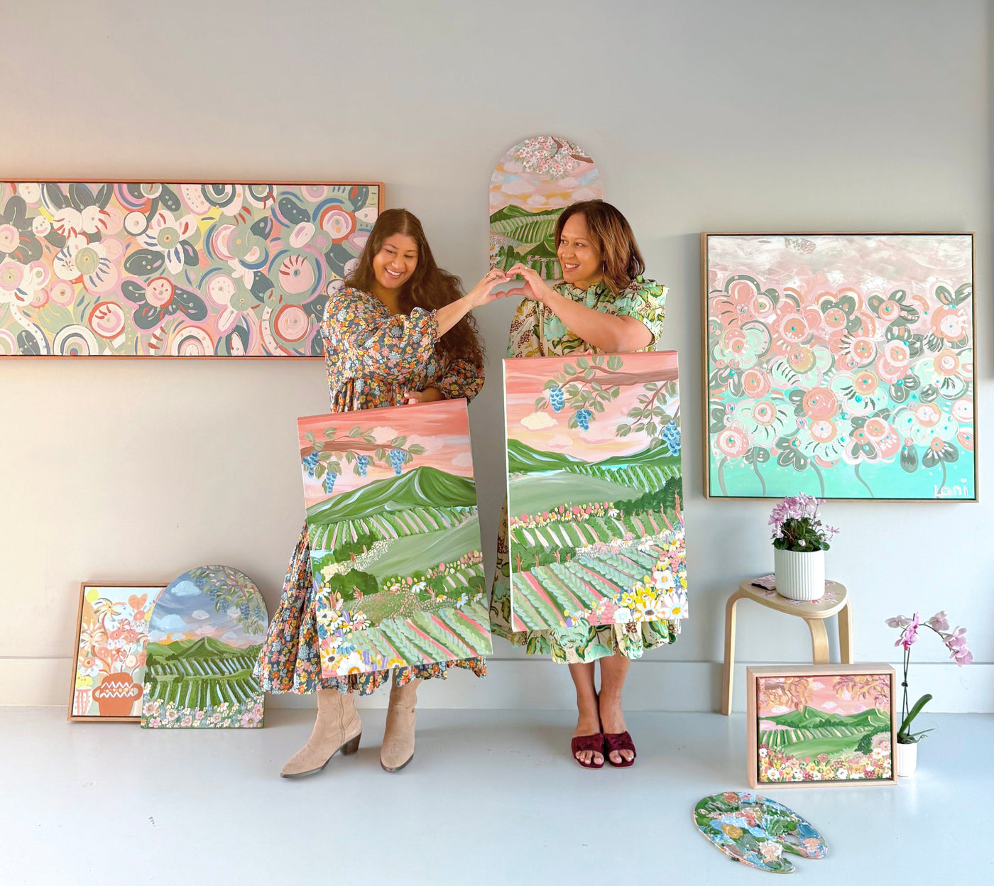 Two women holding a large floral painting in a room with various art pieces on the wall.